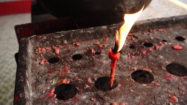 A burning red candle with dripping wax and a bright flame, placed in a rusty, perforated metal holder covered with wax residue, shot in a close-up, high-angle view