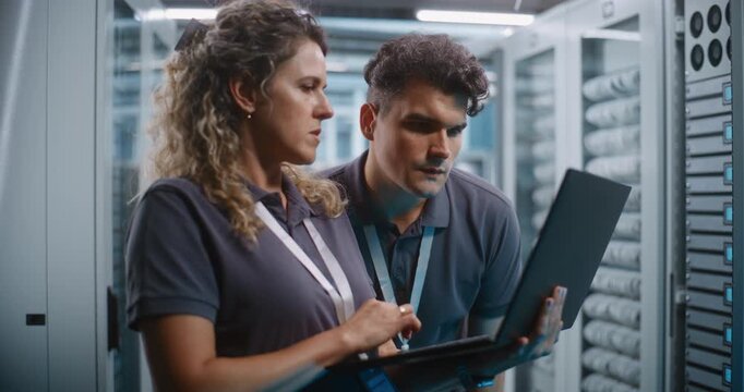 Female IT Specialist Operates Laptop While Teammate Examines Server Rack Hardware Modules in Background. Combined Physical and Software Maintenance Ensures Seamless Cloud Infrastructure Operations.