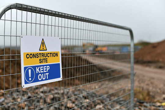 A construction site keep out warning sign is mounted on a metal wire fence in front of an active building area.