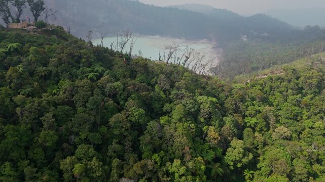 High-angle drone reveal shot flying over a green mountain forest ridge to uncover the misty turquoise volcanic crater lake of Kawah Putih in Ciwidey, Bandung, Indonesia