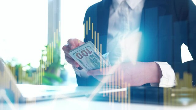 A person is holding a stack of 100 dollar bills in one hand while looking at financial charts on a laptop. Sunlight enters the office, creating a bright environment.
