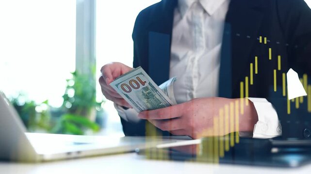 A person is counting cash with a focus on a stack of hundred dollar bills. The person is seated at a desk with a laptop open and financial charts in view. It is daytime in an office.