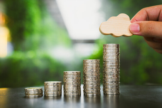 Cloud data storage cost and weather insurance concept with hand placing wooden icon on rising coin stacks on table against green bokeh background for IT infrastructure budget and savings.