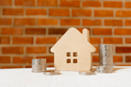 Real estate mortgage and housing savings concept with miniature wooden house model between silver coin stacks on table against brick background for property appraisal and home loan budget.
