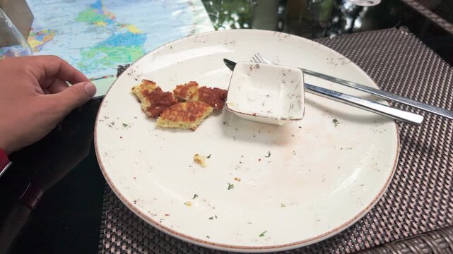 Close-up shot shows a hand next to nearly empty plate with food scraps, utensils, and sauce dish on a woven placemat. Natural daylight, modern outdoor caf&eacute; setting, crisp focus.