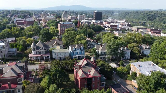 Victorian houses with turrets and porches fill tree lined Lynchburg neighborhood, church spires and downtown high rises rise beyond canopy, distant hills fade in haze. Aerial drone backwards shot