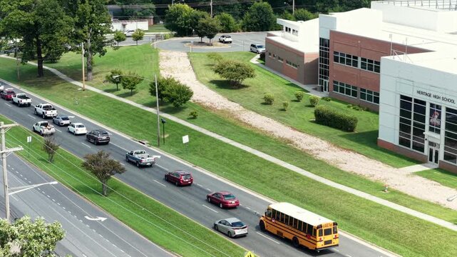 School bus turns onto multilane road beside modern school building, queued trucks and sedans wait behind pedestrian crossing sign, painted arrows mark traffic lanes. Aerial drone tracking shot