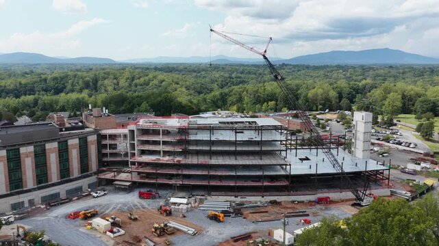 Tower crane lifts materials above multi story steel framework at Lynchburg hospital construction site, exposed concrete floors rise beside completed brick building. Aerial drone rising shot