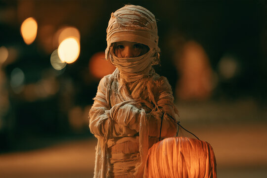 Mummy on Halloween Night Child Holding Glowing Pumpkin Lantern in Spooky Wrapped Costume Outdoors Under Streetlights
