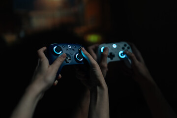 Fototapeta premium Close-up of couple hands holding illuminated wireless game controllers, actively competing in online video game tournament, enjoying leisure time and modern digital entertainment together at home.