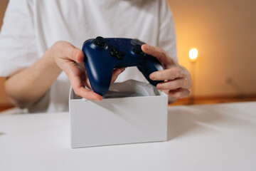 Female hands carefully unboxing new dark blue wireless gamepad controller from white box, revealing modern gaming technology and leisure activity sitting at white table, close-up. © dikushin