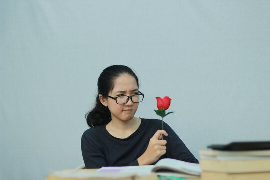 Young woman looking skeptically at a red artificial rose while sitting at a desk, expressing doubt and an unimpressed reaction.