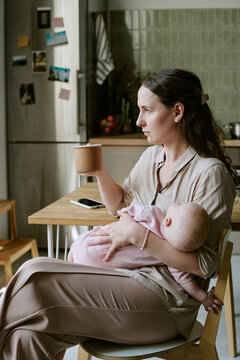 Vertical shot of Caucasian mother holding baby while sitting indoors and drinking from mug. Image suitable for postpartum care campaigns, maternity lifestyle marketing and parenting concepts