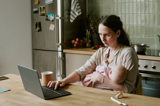 Young adult Caucasian mother holding baby while working on laptop in kitchen. Commercial visual represents maternity leave, remote work challenges, modern parenting lifestyle