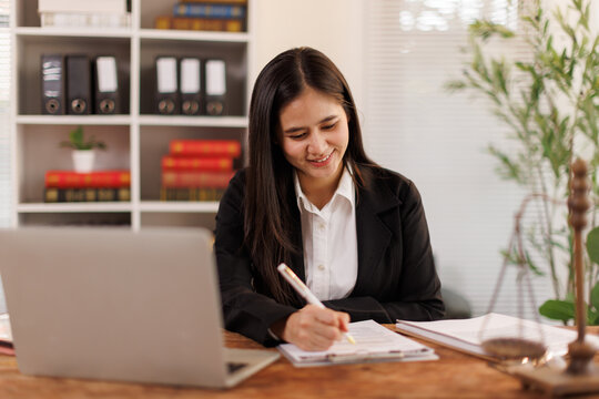 Young legal professional working in law office, reviewing documents and making notes, using laptop for research, scales of justice on desk representing legal system and fairness