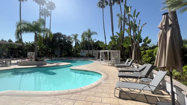 Midday resort swimming pool with empty lounge chairs, tall palm trees and clear blue sky, wide static shot
