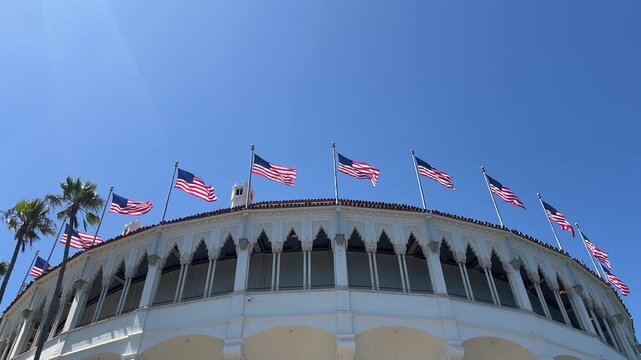 Row of American flags waving above white stucco pavilion with arched windows and palm trees against clear blue sky, low angle daytime shot