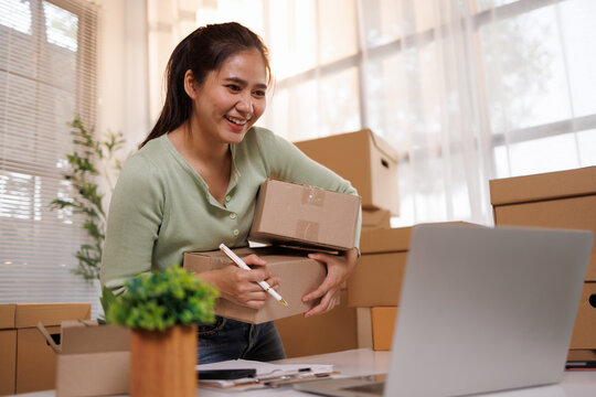 Young asian woman managing her online business, preparing customer orders, holding shipping packages, and using a laptop for e commerce operations in a home office