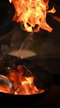 Chef stir-frying meat in a seasoned wok pan with fire flames.  Cinematic vertical video of frying meat in a wok pan in slow motion. Cooking asian cuisine in a restaurant.
