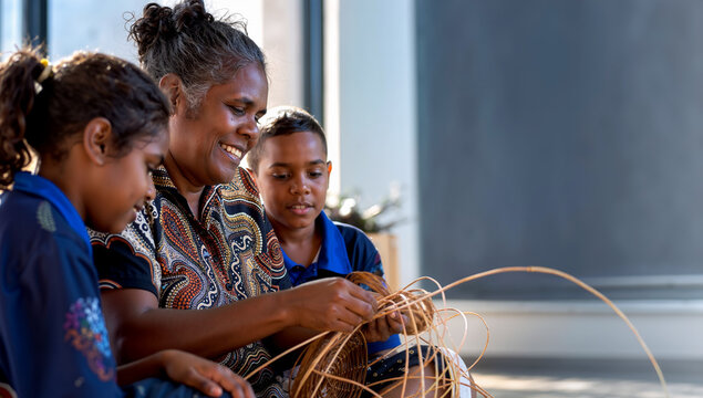 A smiling senior Aboriginal woman teaches two young children the traditional art of basket weaving with natural fibers, sharing indigenous cultural heritage and craft in a bright setting.