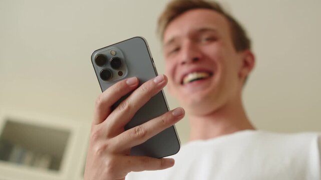 Happy young man using a smartphone indoors. Cheerful young adult male smiling while looking at his mobile phone screen. Man laughing as he is looking into a phone. Watching a funny video.