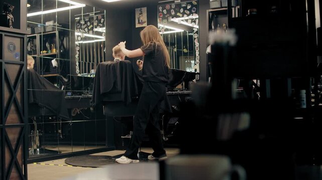 Professional female barber cutting a young boy's hair in a modern barbershop. Small local business. Stylist giving a haircut to a child client in a stylish dark-interior beauty salon. 
