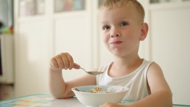 Kid eating organic porridge with a spoon at his small kids table and smiling. Happy little boy sitting at a table and eating hot porridge for breakfast at home. 