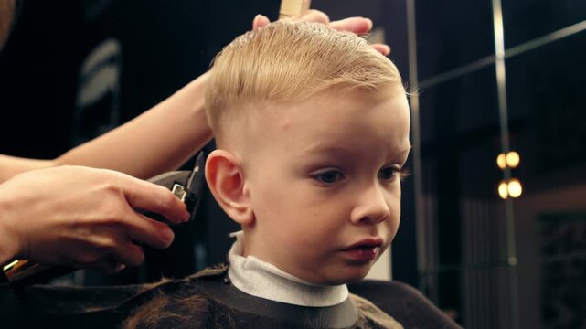 Professional female barber cutting a young boy's hair in a modern barbershop with a trimmer. Stylist trimming a child in a stylish beauty salon with a buzzer.
