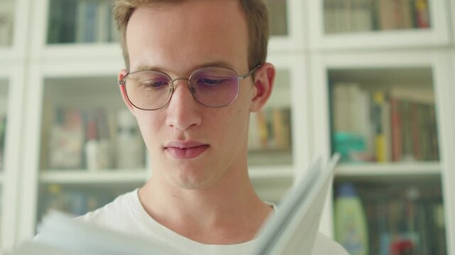 Young man with glasses reading a book in a library. Smart teen studying in a bookstore. University student in eyeglasses reading a paperback book in a library. Portrait of a young adult flipping pages