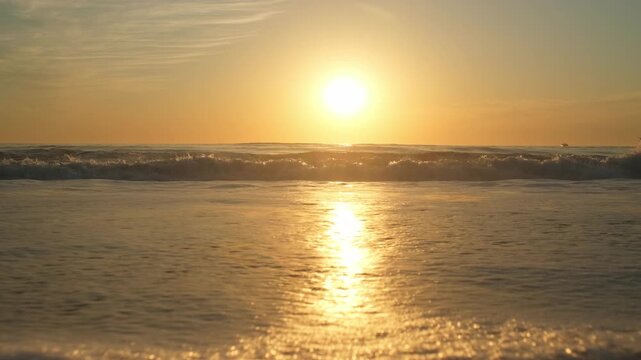 Low angle view of  Ocean shore with rolling waves with an orange sun on the horizon while sunset. Sandy beach with rolling waves during sunset in summer. Contemplation of the sea waves during sunset.