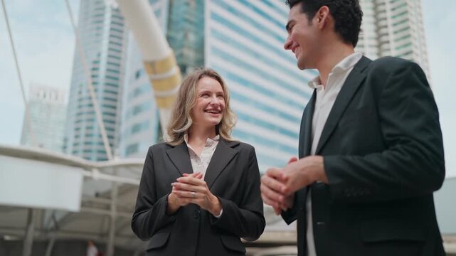 Two professionals converse near modern skyscrapers. They smile, showing friendly rapport. Business colleagues stroll through urban setting