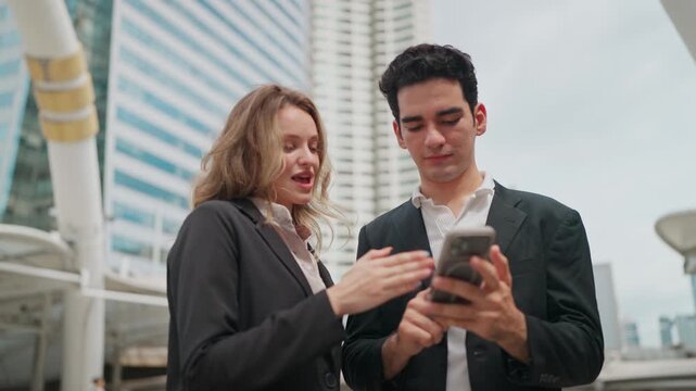 Business couple checks phone together outdoors. They smile, focused on shared screen content. Urban backdrop suggests professional collaboration