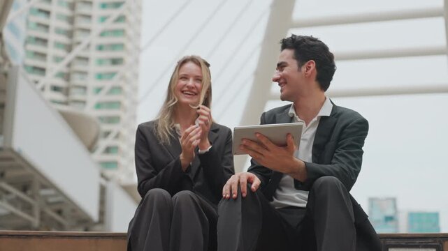 Two professionals share a tablet outdoors, smiling together. They sit on a modern structure, surrounded by tall buildings