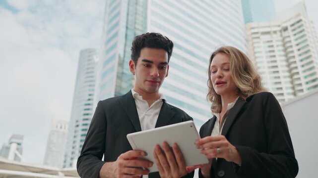 Business professionals discuss plans using a tablet outdoors. They stand near tall buildings, dressed in suits