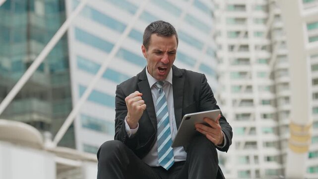 Businessman sits outside modern building. He grips tablet, showing excitement. He clenches fist, smiling broadly