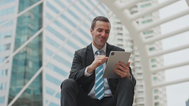 Businessman sits thoughtfully outdoors. He holds tablet, dressed smartly. Urban towers loom behind him. He looks upward, smiling slightly
