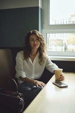 A woman with curly hair sits at a wooden table in a cafe, holding a paper cup. Sunlight streams through the window, illuminating her casual outfit as she seems relaxed and content