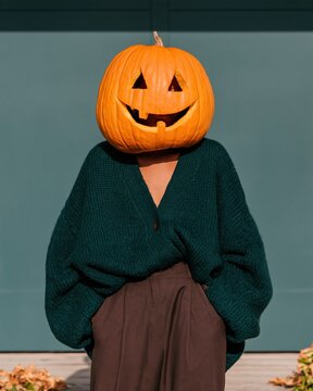 Carved pumpkin head on a woman wearing a cozy teal cardigan and charcoal trousers on a porch, for surreal halloween and autumn themes