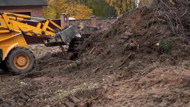 Yellow loader moves through muddy pile. Tire tracks mark wet ground. Bare trees stand behind fence. Worker operates machine. Effort is visible in each motion