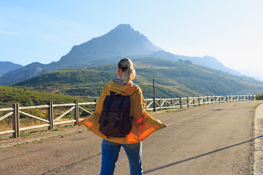 Woman backpacker standing on a winding road, admiring the vast mountain range and clear sky, feeling freedom and adventure while traveling through scenic nature