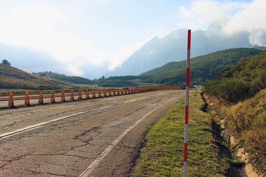 Empty asphalt road curves through a mountain pass with snow pole and safety cones in foreground, misty peaks under cloudy sky evoking a remote scenic journey in picos de europa