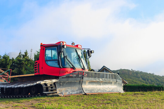 Red snowcat groomer parked on green grass, awaiting winter operations, with forest trees and mountain landscape under a bright sky