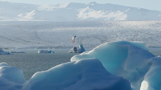 Kitesurfer rides among icebergs in Jokulsarlon glacier lagoon beneath Breidamerkurjokull, with the Vatnajokull ice cap and rugged mountains beyond in Hornafjordur, Iceland, aerial slide