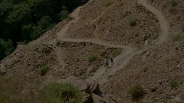 High-angle view of a traveler riding a mule guided by a local on a winding mountain trail in Morocco. Winding dirt path cut into dry rocky hillside terrain.