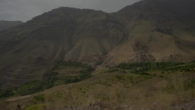 Panoramic wide shot of massive High Atlas mountain ridges towering over stepped agricultural terraces, winding switchback roads, and a Berber settlement in Morocco.