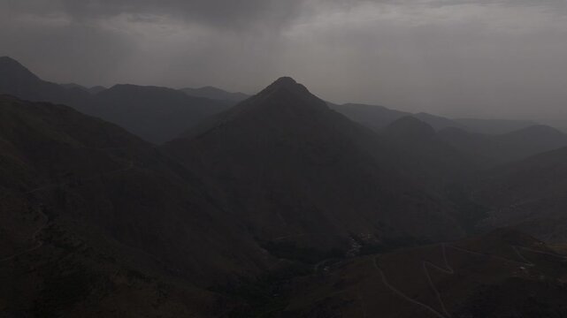 Dramatic aerial wide shot of a sharp mountain peak rising over deep valleys and switchback roads under dark, stormy skies in Morocco's High Atlas.
