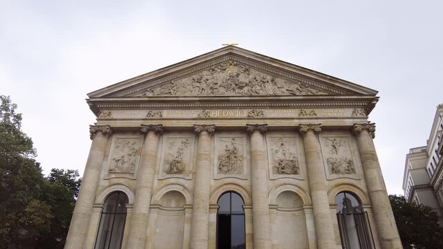 approach toward St. Hedwig's Cathedral reveals detailed pediment sculptures and Corinthian columns under overcast skies.