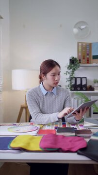 Young Asian fashion designer reviews color swatches with tablet in bright home studio workspace