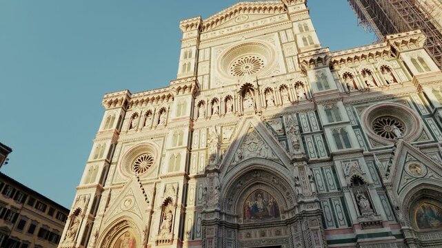 The polychrome marble front of Santa Maria del Fiore rises above the square with its gabled portal, mosaics and rose window in Florence, Tuscany. Ground level tilt up the facade, low angle.