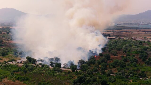 A mass of smoke has settled over the fields inland of Olbia, Sardinia, leaving only the Gallura ridgelines legible above it. Aerial view, rising camera.